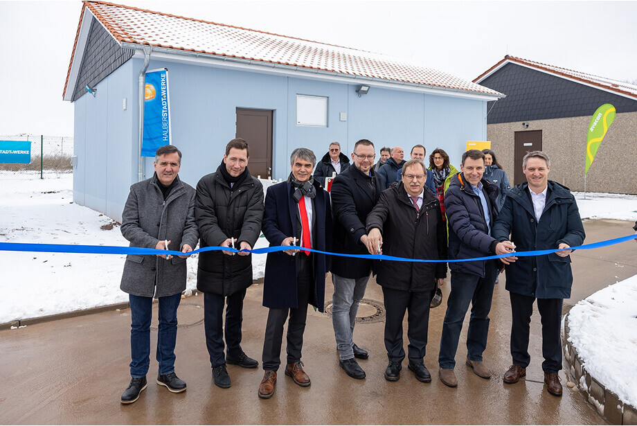 Im Vordergrund durchschneiden Herr Balcerowski, Herr Szarata, Herr Dr. Eichner,  Herr Abram-Hohnl, Herr Prof. Dr. Gerloff, Herr Heinrich und Herr Ereth ein blaues Band. Im Hintergrund ist das neue Schalthaus in blau zu sehen. Hinten links steht eine Beachflag der Halberstadtwerke. Es liegt Schnee.