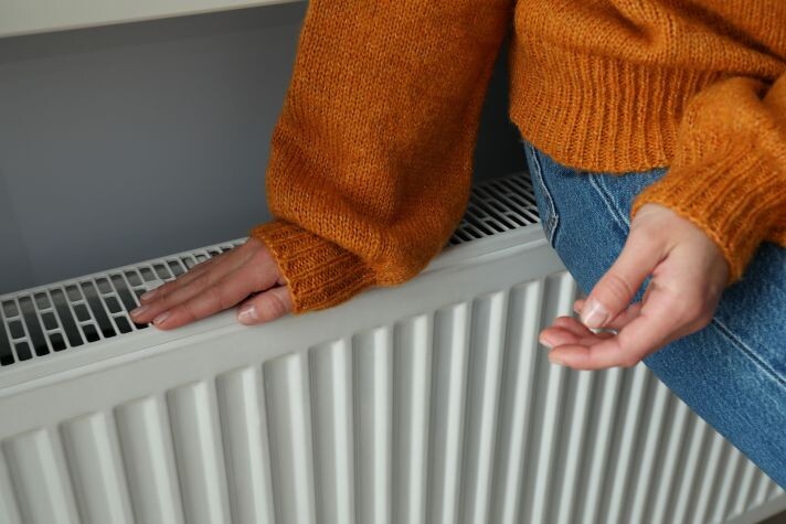 Eine Frau in Jeanshose und orangenem Strickpullover sitzt auf einem Heizkörper. Eine Hand legt sie dabei prüfend auf die Abdeckung des Heizkörpers.
