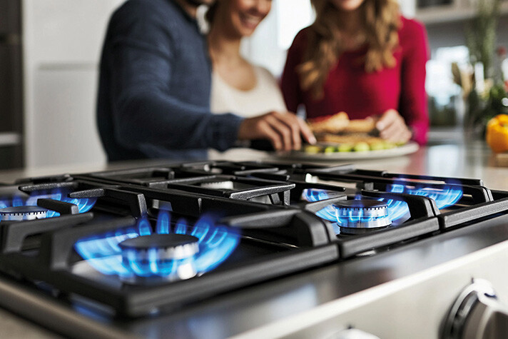 Drei Personen bereiten in einer Küche augenscheinlich das Essen vor – im Vordergrund brennen bereits die Blauen Flammen eines eingeschalteten Gaskochfeldes.