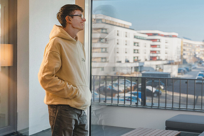 Ein junger Mann lehnt sich in seiner Wohnung lässig gegen seine geschlossene Balkontür und genießt augenscheinlich die wohlige Wärme im Zimmer. Seine Wohneinheit befindet sich im Gebäude-Ensemble der Halberstädter Lindenhof-Terrassen, eines der größten Anschlussobjekte am hiesigen Fernwärme-Netz - dieses ist teilweise in der Hintergrundansicht des Balkons erkennbar. 