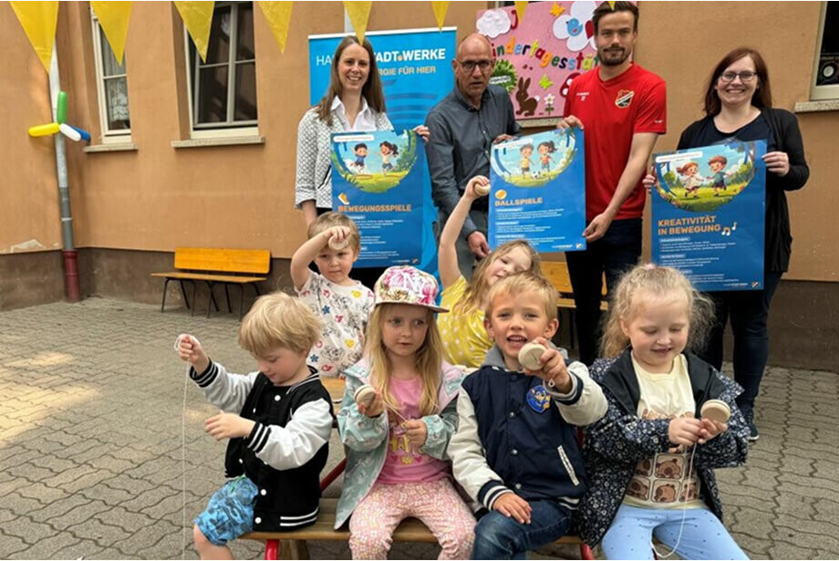 Eine Gruppe von Kindern sitzt mit Holz-Jojos auf Bänken vor einem Gebäude. Im Hintergrund halten Frau Pinkernelle, Herr Hartmann, Herr Guderitz und Frau Lücke drei Plakate in die Kamera. Hinten links ist ein Roll-Up der Halberstadtwerke, sowie eine gelbe Wimpelkette und bunte Luftballons zu sehen.