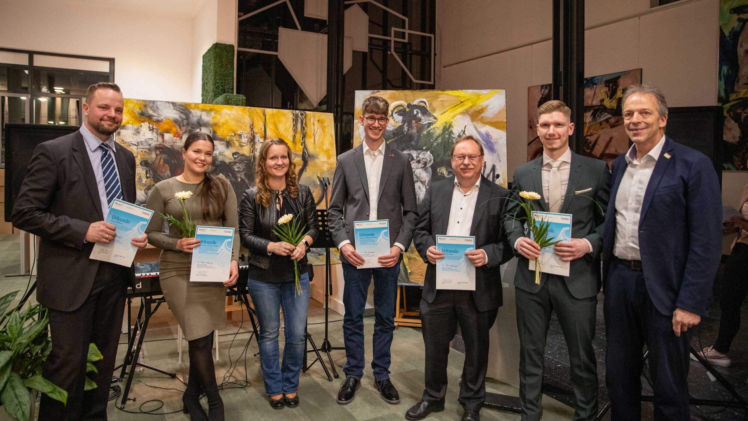 Eine festlich angezogene Gruppe von Menschen verschiedenen Alters steht nebeneinander im Halbkreis auf einer Bühne. Fünf von ihnen halten eine Urkunde in der Hand, drei von ihnen eine gelbe Gerbera. Ganz rechts stehen Prof. Dr. Gerloff und Prof. Dr. Roland. Im Hintergrund sieht man zahlreiche große und kleine Gemälde.