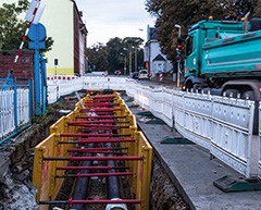 Auf einer Baustelle werden neue Rohre verlegt. Im Vordergrund dieneue Rohre in einer gelben Einfassung. Rechts daneben ein türkisfarbenes Baustellenfahrzeug hinter einer weißen Absperrung.