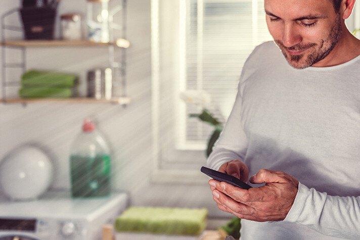 Ein Mann in weissem Langarmshirt steht in einem Hauswirtschaftsraum und nutzt vertieft sein Smartphone – hinter ihm läuft gerade eine Waschmaschine und die Sonne scheint durch Jalousie des Raumes.