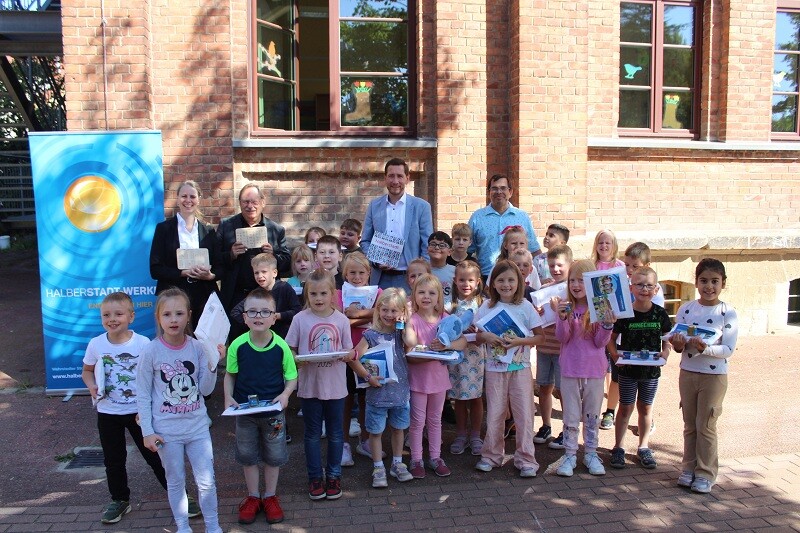 Eine Gruppe von Kindern steht mit einem Halberstadtwerke-Umschlag zusammen mit Herrn Lütgert, Frau Pinkernelle, Herrn Prof. Dr. Gerloff und Herrn Szarata im Schatten vor einem Backstein-Gebäude. Hinten links sieht man ein Roll-Up der Halberstadtwerke.