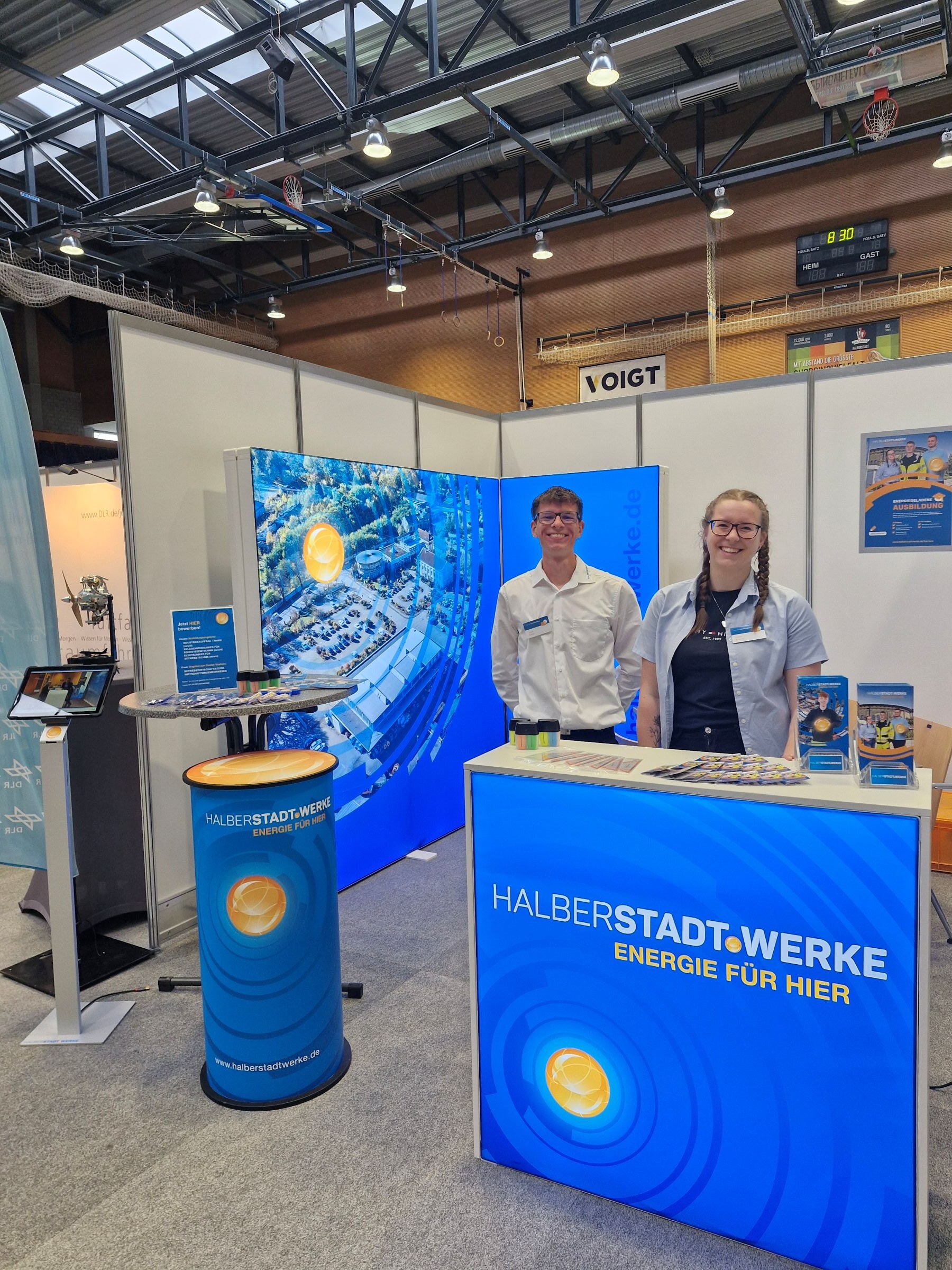 Ein junger Mitarbeiter und eine junge Mitarbeiterin der Halberstadtwerke stehen zusammen an dem Messestand des Unternehmens in einer Messehalle.