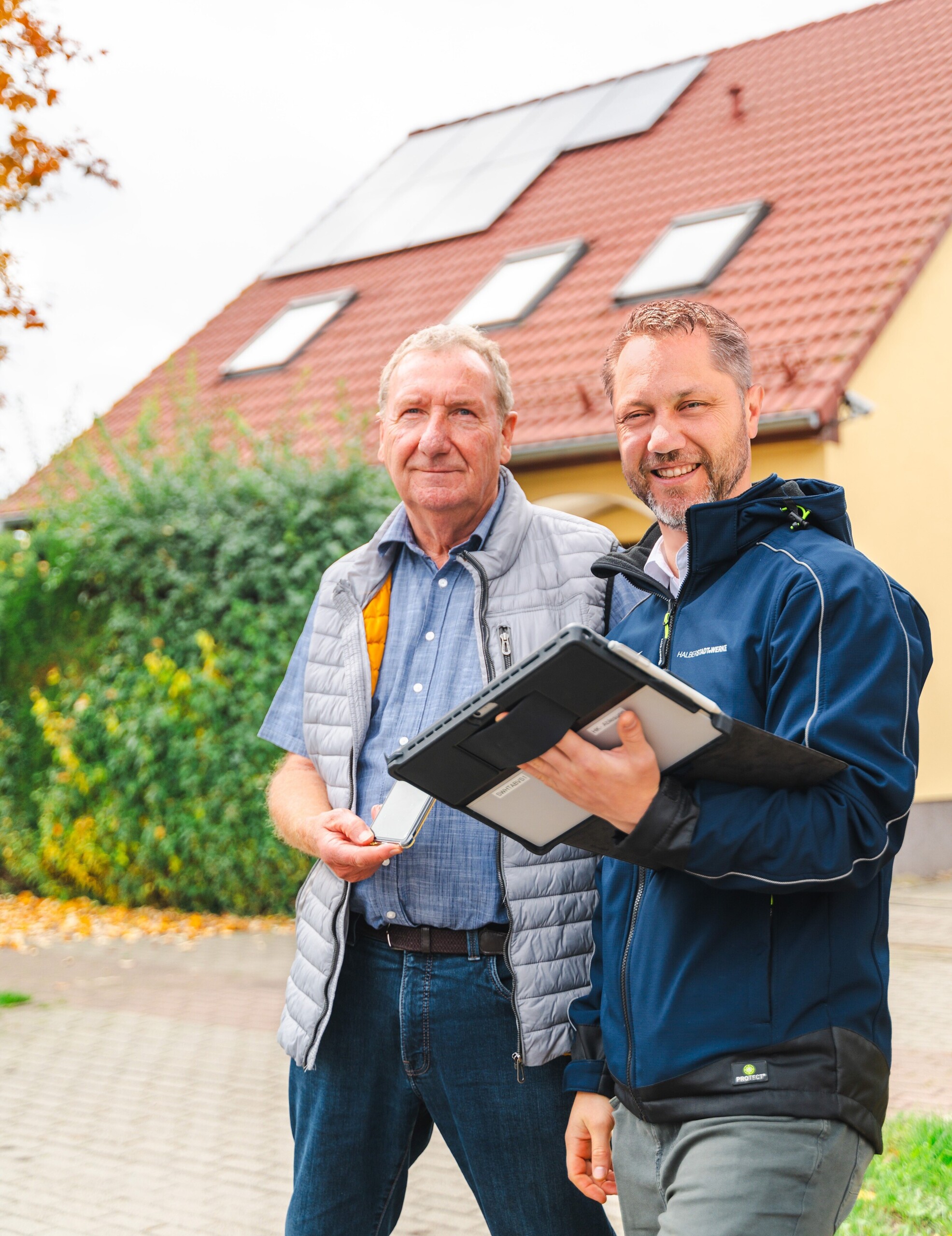 Horst Cernota, PV-Kunde der Halberstadtwerke steht neben Nico Ihsecke, Projektmanager PV der Halberstadtwerke. Beide schauen lächeln in die Kamera. Im Hintergrund ist das gelbe Einfamilienhaus von Herrn Cernota zu sehen.