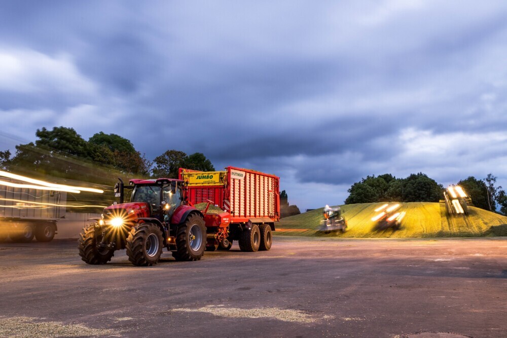 Einbringen der Maisernte auf dem Gelände der Biogasanlage in der Abenddämmerung. Im Vordergrund fährt ein roter, leerer Traktor mit Anhänger und mit Scheinwerferlicht gerade davon. Im Hintergrund fahren drei Fahrzeuge im Zeitraffer-Augenblick auf den ansteigenden Silage-Hügel.